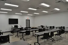 Thumbnail for Empty modern classroom with black chairs, gray tables, wall-mounted screen, and fluorescent ceiling lights.