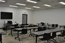 Thumbnail for Modern empty classroom with black chairs, rectangular tables, a large screen, and white walls under fluorescent lights.