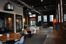 Thumbnail for Modern industrial office space with round wooden tables, white chairs, exposed brick walls, and large windows.