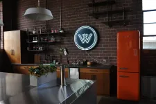 Thumbnail for Industrial style kitchen with brick walls, neon sign, orange retro fridge, wooden cabinets, and modern stainless steel island.