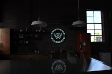 Thumbnail for Dark industrial kitchen with brick walls, hanging lights, neon W logo, and orange refrigerator by a window.