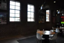 Thumbnail for Cozy cafe interior with round wooden tables, white chairs, large windows, and industrial pendant lights.