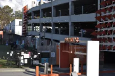 Thumbnail for Outdoor parking garage entrance with orange signage and nearby green fenced area under partly cloudy sky.