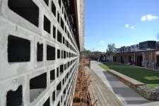 Thumbnail for Modern outdoor space with patterned concrete wall, wooden benches, green lawn, and urban buildings under blue sky.