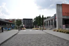 Thumbnail for Wide pedestrian plaza with modern commercial buildings, trees, and textured pavement under partly cloudy skies.