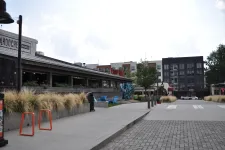 Thumbnail for Outdoor urban scene with modern buildings, a restaurant patio, ornamental grasses, and pedestrian pathways under a partly cloudy sky.