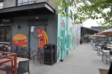 Thumbnail for Outdoor seating area with mural of cartoon burger and hot dog high-fiving on brick wall at restaurant.