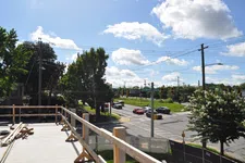 Thumbnail for View of a sunny suburban intersection with cars, trees, and construction site railing in the foreground.