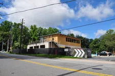 Thumbnail for Two-story building under construction with wooden framing, black fencing, and clear blue sky with clouds.