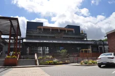 Thumbnail for Modern two-story restaurant building with outdoor patios, greenery, and parked white car under a partly cloudy sky.