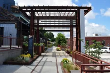 Thumbnail for Modern outdoor walkway with metal pergola, planters, and urban buildings under a blue sky with clouds.