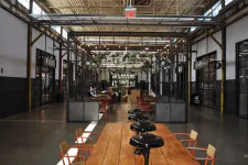 Thumbnail for Open industrial-style coworking space with wooden tables, orange chairs, hanging plants, and exposed ceiling lighting.