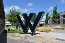 Thumbnail for Large black W sculpture with blue graffiti in an outdoor plaza with green chairs and trees under a blue sky