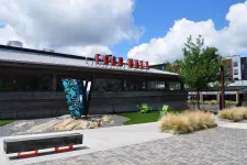 Thumbnail for Outdoor view of a modern food hall with wooden facade, red sign, green chairs, and landscaped plaza under blue sky.