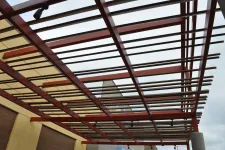 Thumbnail for Modern pergola with wooden slats and red beams over a building exterior under cloudy sky.
