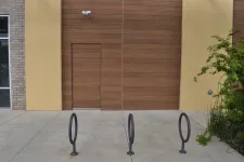 Thumbnail for Empty bike racks in front of a modern wooden and yellow building wall with a concealed door and greenery.