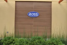 Thumbnail for Blue Ross store logo mounted on wooden panel wall with green plants in front under beige structure.