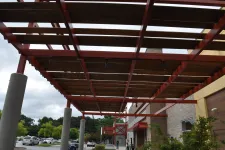 Thumbnail for Outdoor walkway with red steel pergola and wooden slats next to a brick building under cloudy sky.