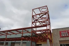 Thumbnail for Red metal framework structure with circular cutouts above a building entrance under cloudy sky.