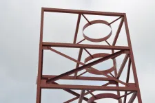 Thumbnail for Rust-colored metal framework with circular and diagonal supports against a cloudy sky.