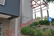 Thumbnail for Outdoor staircase with red railings next to modern building and green shrubbery under metal canopy.