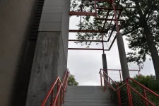 Thumbnail for Concrete stairs with red railings lead up to a metal frame structure surrounded by trees against a cloudy sky.