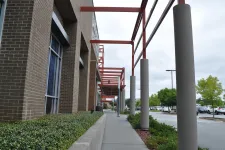 Thumbnail for Sidewalk beside a brick building with red steel beams and concrete pillars under an overcast sky near a parking lot.