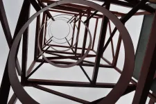 Thumbnail for Looking upward inside a red metal tower structure with circular and diagonal beams against a cloudy sky.