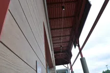 Thumbnail for View of angled building exterior with beige siding and brown metal roof structure against cloudy sky.