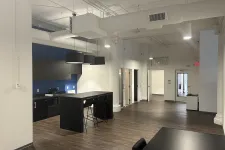 Thumbnail for Modern office kitchen area with black island, bar stools, blue accent wall, and open hallway with wood floors.