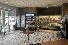Thumbnail for Interior of a deli with refrigerated display cases, shelves of food products, and a central table with baked goods.