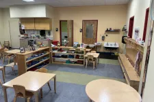 Thumbnail for Empty preschool classroom with small wooden tables, chairs, shelves, and art supplies neatly organized.