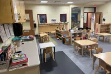 Thumbnail for Bright preschool classroom with small wooden tables, chairs, shelves, and educational materials.