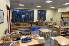 Thumbnail for Montessori classroom with wooden tables, chairs, educational materials, and large windows letting in natural light