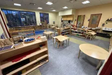Thumbnail for Spacious Montessori classroom with wooden tables, chairs, learning materials, and large windows for natural light.