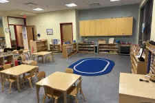 Thumbnail for Bright and organized classroom with wooden tables, chairs, shelves, and a blue oval rug on gray floor.