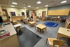Thumbnail for Spacious preschool classroom with small tables, chairs, shelves, and a blue carpet area for group activities.