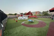 Thumbnail for Empty modern playground with artificial turf, red and gray play elements, and a shaded canopy near buildings.