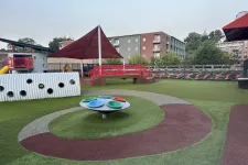 Thumbnail for Modern playground with colorful round play panels, shade canopy, and soft turf in an urban setting.