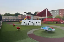 Thumbnail for Modern outdoor playground with colorful spinning discs, slide, climbing structures, and shaded red canopy over seating area.