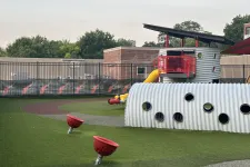 Thumbnail for Modern playground with metal tunnel, red spinning seats, yellow slide, and turf surface under overcast sky