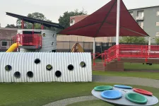 Thumbnail for Modern playground with a large red shade canopy, corrugated tunnel, colorful spinning play equipment, and a slide.