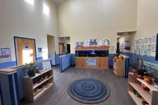 Thumbnail for Bright, spacious daycare lobby with blue circular rug, fish tank, shelves with toys, and children's artwork on walls.