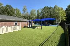Thumbnail for Outdoor playground with artificial turf, blue shade canopies, slides, and a brick building surrounded by trees.