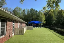 Thumbnail for Outdoor playground with artificial turf, blue shade structure, and brick building under clear blue sky.