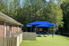 Thumbnail for Outdoor playground with blue shade sails, artificial grass, play structures, and fenced area next to brick building.