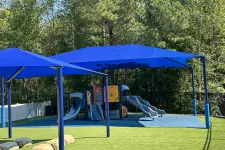 Thumbnail for Outdoor playground with blue slides, climbing structures, and blue shade canopies surrounded by greenery.