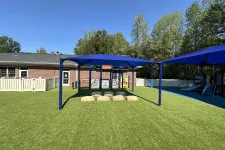Thumbnail for Playground area with artificial grass, blue shaded canopies, stepping stones, and a brick building in background