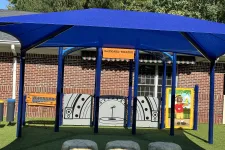 Thumbnail for Outdoor children's play theater area with blue canopy, stage backdrop, and artificial grass seating resembling rocks.