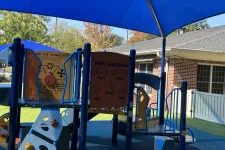 Thumbnail for Children's playground with sign language learning panel and colorful climbing structures under a blue shade canopy.